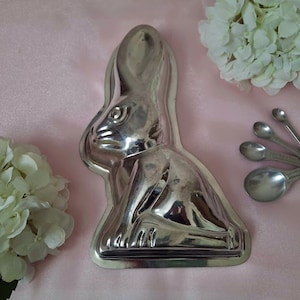 May include: A silver rabbit-shaped mold for baking chocolate or other treats. The mold is on a pink surface with white flowers.