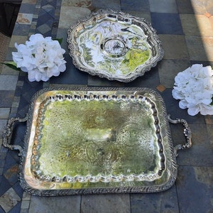 May include: Two ornate silver serving trays. One is rectangular with handles, the other is round with a scalloped edge. White flowers are placed near the trays. The trays have intricate embossed designs and reflect the surrounding environment.