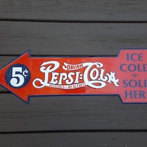 May include: Vintage Pepsi-Cola arrow-shaped sign. The sign is red with white and blue lettering. It reads "Drink Pepsi-Cola" and "Ice Cold Sold Here." The sign also features the price "5&cent;".