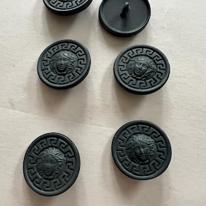 Puede incluir: Cinco botones negros con un diseño en relieve de una cara de mujer rodeada por un patrón de clave griega.