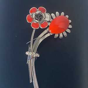 May include: A silver brooch with two orange enamel flowers. One flower is a rose with a black center and the other is a stylized flower with a large orange oval in the center. The brooch has a cluster of clear rhinestones on the stem.