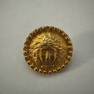 May include: A circular gold-toned button featuring a raised relief of a face with flowing hair in the center. The edge of the button is decorated with a series of small, evenly spaced raised dots that form a border around the central design.