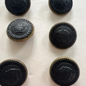 Puede incluir: Seis botones negros y dorados con un diseño en relieve de una cara. Los botones tienen un borde dorado y un centro negro con un diseño en relieve de una cara.