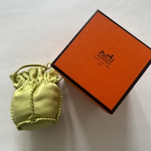 May include: A small, light green leather pouch with a drawstring closure. The pouch is sitting on a white surface next to an orange box with the Hermes logo and the words "Hermes, Paris" printed on it.