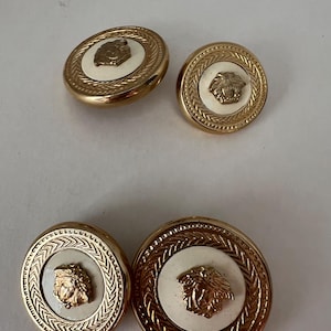 Peut inclure: Quatre boutons dorés avec un centre blanc et un motif de tête de lion doré. Les boutons ont une bordure dorée décorative.