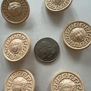 May include: Six beige round buttons with a raised gold design of a face with a Greek key border. The buttons are approximately the size of a US quarter.