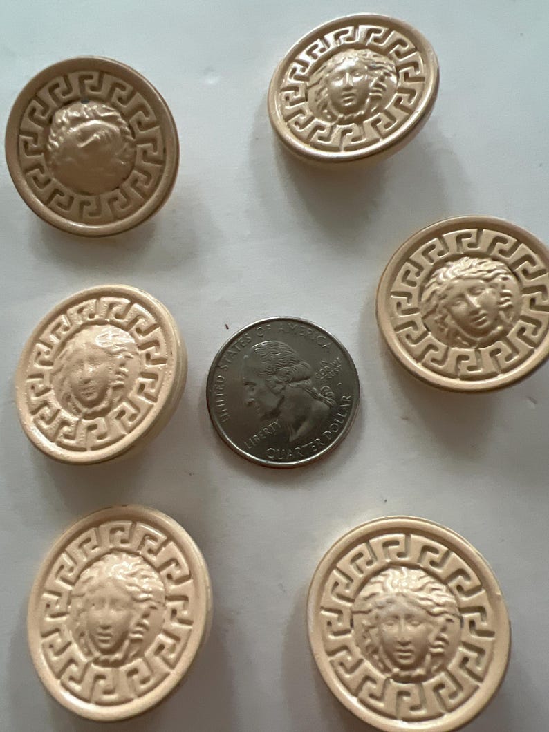 May include: Six beige round buttons with a raised gold design of a face with a Greek key border. The buttons are approximately the size of a US quarter.