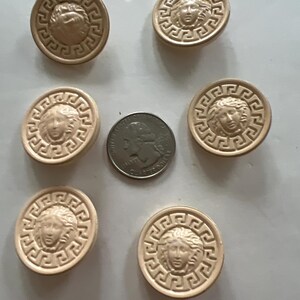 May include: Six beige round buttons with a raised design of a woman's face surrounded by a Greek key pattern.