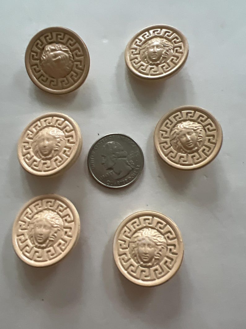 May include: Six beige round buttons with a raised design of a woman's face surrounded by a Greek key pattern.