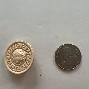 May include: A beige ceramic button with a raised design of a woman's face surrounded by a Greek key pattern. The button is next to a US quarter.