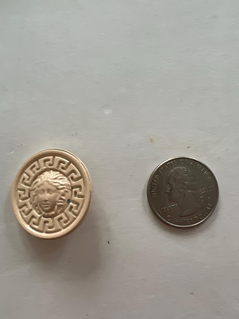 May include: A beige ceramic button with a raised design of a woman's face surrounded by a Greek key pattern. The button is next to a US quarter.
