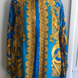 May include: A long-sleeved button-down shirt with a blue background and a gold baroque pattern. The shirt has a collar and a button-up front.