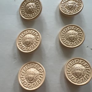 May include: Six beige round buttons with a raised design of a face surrounded by a Greek key pattern.