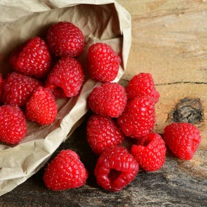 May include: A collection of fresh, bright red raspberries spilling out of a crumpled brown paper bag. The berries feature a textured, bumpy surface and are scattered across the frame, showcasing their vibrant color and rounded, hollow-centered shapes.
