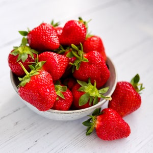 Puede incluir: Un cuenco de cerámica lleno de fresas frescas de color rojo brillante, cada una con un tallo verde con hojas. Varias fresas están agrupadas dentro del cuenco, mientras que otras dos fresas descansan fuera del cuenco sobre la superficie.