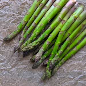 May include: Fresh green asparagus spears with purple tips are arranged on a crumpled brown paper surface. The spears are vibrant green, with a slightly purple hue at the tips. The image is a close-up shot.