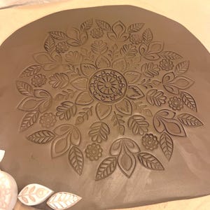 May include: A brown clay slab with an embossed floral design. The design features a central circular element surrounded by leaves and flowers. The details are finely carved, creating a textured surface.