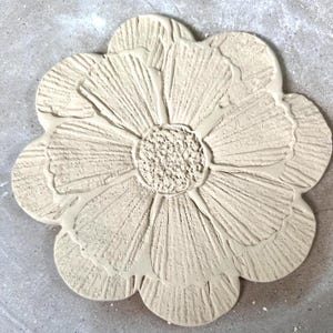 May include: A circular, light beige clay relief sculpture shaped like a flower with ten distinct petals. The petals feature a textured, ribbed surface pattern radiating from a central, densely stippled circular core, creating a tactile finish.