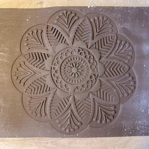 May include: A brown clay tile with an intricate, embossed mandala design. The mandala features a central circle surrounded by eight symmetrical petals with detailed patterns.