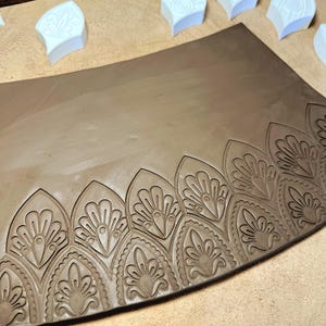 May include: A flat, rectangular slab of brown clay featuring a repeating embossed pattern of pointed, petal-like arches along the bottom edge. Several white, pointed plastic stamps with matching designs are positioned above the clay slab.