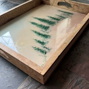May include: Wooden serving tray with a light-colored resin surface. The resin features a forest scene with dark green trees. The tray has a rectangular shape with cut-out handles on the sides.