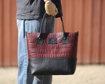 May include: A burgundy leather tote bag with a black leather bottom and crocodile embossed pattern. The bag has two black leather handles and a brass buckle on each side.