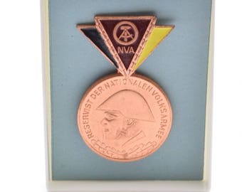 May include: A bronze medal with a profile of a person wearing a military hat. The medal is surrounded by a wreath and the text "Reservist der Nationalen Volksarmee". The medal is displayed in a white and blue box with a red, black, and yellow triangle with the text "NVA" in the center.