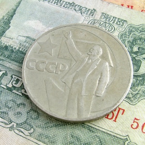 Può includere: Una moneta d'argento con un'immagine in rilievo di un uomo che alza il braccio in aria. La moneta ha il testo "CCCP", che è l'abbreviazione cirillica per l'Unione delle Repubbliche Socialiste Sovietiche. La moneta è appoggiata su una banconota verde e bianca con il numero "3" stampato su di essa.