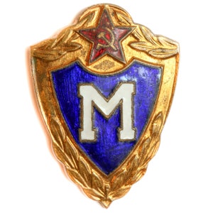 Puede incluir: Un pin de metal dorado con un escudo azul que presenta una letra "M" blanca. El escudo está rodeado por una corona de hojas doradas y una estrella roja con un martillo y una hoz.