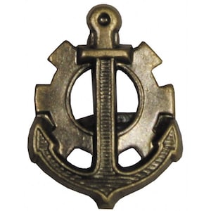 May include: Bronze-colored metal pin featuring an anchor design overlaid on a gear. The anchor's central shaft is vertically oriented, with a curved base. The gear's teeth are visible on either side of the anchor, and the pin has a circular opening.