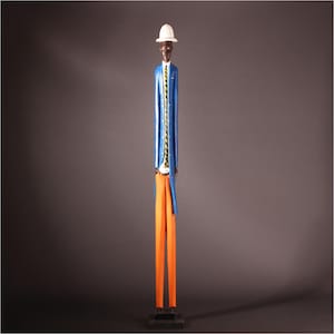 Op de afbeelding: Een lange, slanke houten figuur van een persoon met een witte hoed, blauwe jas, oranje broek en zwarte schoenen. De jas heeft een geel en zwart touwachtig detail. De figuur staat op een zwarte basis tegen een donkergrijze achtergrond.
