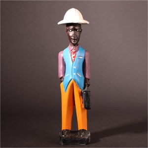 May include: A carved wooden figurine of a person wearing a white hat, purple shirt, blue vest, orange pants, and black shoes. The figurine is holding a black briefcase. The figure is standing on a black base.