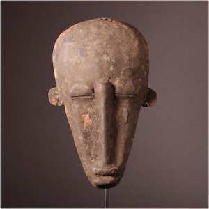 Puede incluir: Una máscara de madera con una cara larga y estrecha y los ojos cerrados. La máscara está tallada en un estilo sencillo y tiene una pátina marrón oscura.