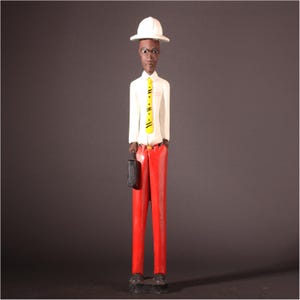 May include: A carved wooden figurine of a person in a white shirt, yellow tie, and red pants. The figurine is holding a black briefcase and wearing a white hard hat. The figure is standing on a black base.