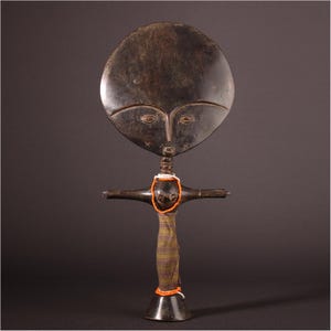 May include: A dark wooden sculpture of a stylized figure with a large, round head and a cross-shaped body. The figure has carved facial features and is adorned with beaded necklaces and a wrapped fabric skirt. The sculpture is a unique piece of art.