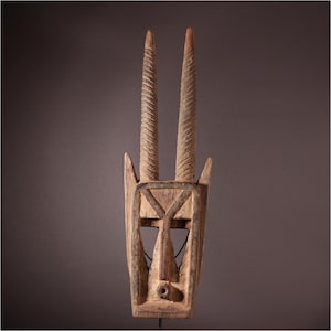 May include: A carved wooden mask with two long, spiraled horns. The mask has a rectangular face with geometric cutouts and a small, round nose. The wood has a natural, aged appearance, with visible grain and variations in color.