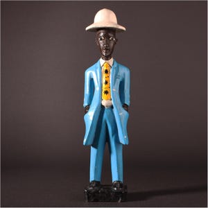 May include: A carved wooden figurine of a man in a light blue suit, white hat, and yellow tie with a star pattern. The figurine has black shoes and is standing on a black base. The figure's skin is dark brown.