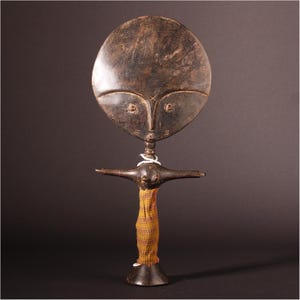 May include: A wooden sculpture of a stylized female figure with a large circular head and outstretched arms. The figure is painted in brown and black, and has a yellow and brown fabric wrapped around its torso.