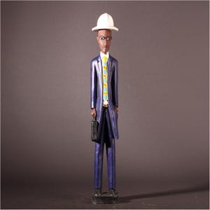 May include: A carved wooden figurine of a man in a blue suit, white hat, and yellow tie with blue accents. The figurine is holding a black briefcase and stands on a black base. The figure has dark skin and is painted with fine details.