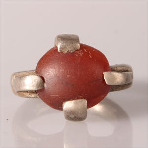 May include: A silver ring with a red carnelian stone set in the center. The stone is surrounded by four silver prongs.