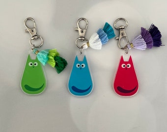 May include: Three keychains with colorful cat charms. Each charm is a different color: green, blue, and red. The keychains have colorful tassels.