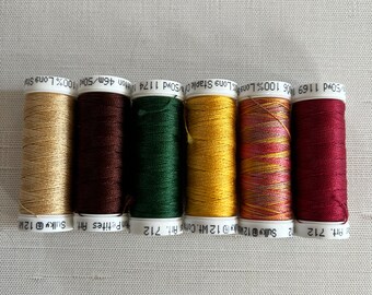 May include: A row of six spools of thread in various colors, including gold, burgundy, dark green, yellow, and red. The spools are labeled with text indicating the thread type and length. The threads are likely for sewing or embroidery.