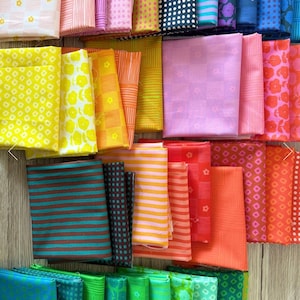May include: A selection of folded fabric squares in a variety of colours and patterns. The fabrics include yellow with floral designs, orange with stripes, pink with small flowers, and teal with brown stripes. Other colours include red, green, and blue.