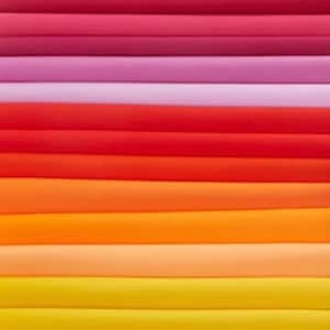 Puede incluir: Primer plano de una pila de tela en degradado de colores. Los colores transitan desde rojos y rosas intensos en la parte superior, pasando por tonos de naranja y amarillo en la parte inferior. La tela parece lisa y de tejido apretado.