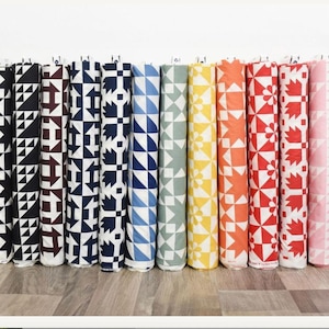 Pode incluir: Uma coleção de rolos de tecido com vários padrões geométricos. Os desenhos incluem triângulos, quadrados e formas de folhas em cores como preto, azul marinho, bordô, azul, verde, amarelo, laranja, vermelho e rosa. Os tecidos estão dispostos numa prateleira branca.