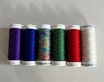 May include: Six spools of colorful embroidery thread in shades of purple, blue, multicolored, green, red, and off-white. Each spool has a white label with black text. The threads are arranged horizontally.