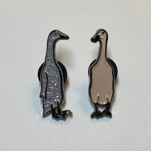 May include: Two enamel pins featuring a goose design. The goose on the left is silver with glitter, while the goose on the right is beige.