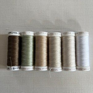 Könnte beinhalten: Sechs Garnrollen in einer Reihe, in verschiedenen neutralen Farben wie Braun, Grün, Beige und Weiß. Die Spulen sind mit Informationen über Garnart und -länge beschriftet, jede enthält etwa 46 Meter Garn.