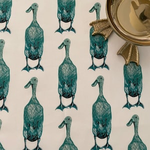 Pode incluir: Tecido branco com um padrão de patos verde-azulado. Uma tigela de metal dourado com alças em forma de pés de pato está no canto superior direito. Os patos são estilizados com pescoços longos e pés palmados.