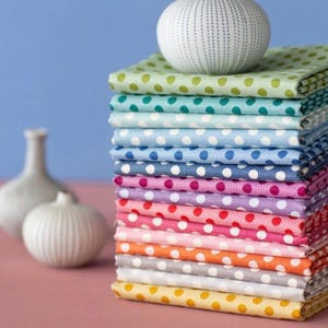 Pode incluir: Uma pilha de quadrados de tecido coloridos com bolinhas brancas. Os tecidos são de várias cores, incluindo verde, azul, rosa e laranja. Uma esfera branca texturizada está no topo da pilha. Vasos brancos decorativos no fundo.
